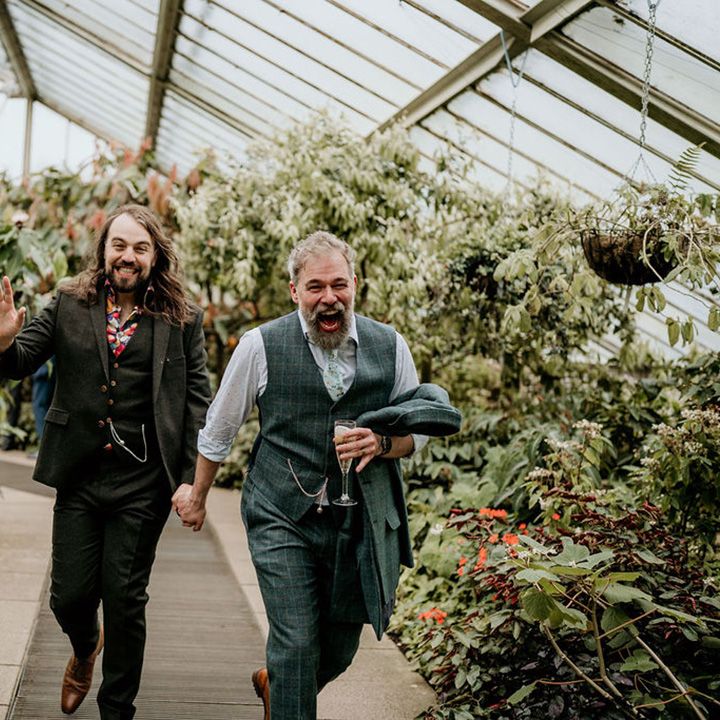 Grooms laugh and run through botanical greenhouse filled with lush greenery and florals at Kew Gardens 