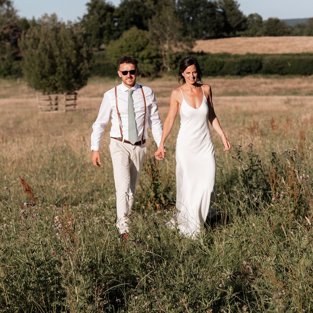 Bride in silk Grace Loves Lace wedding dress walks alongside her groom in white shirt and braces with sage green tie 
