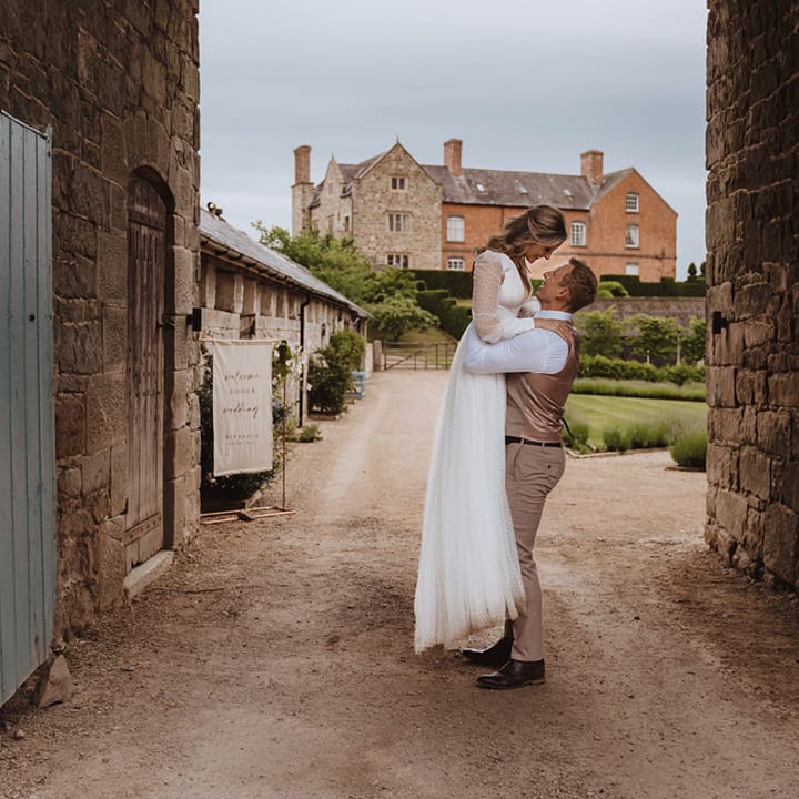 The groom in a beige waistcoat and trousers lifts the bride in a long sleeve wedding dress up in the air 