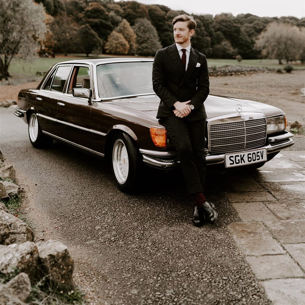 The groom in a pinstriped suit standing against the brown Mercedes wedding car 