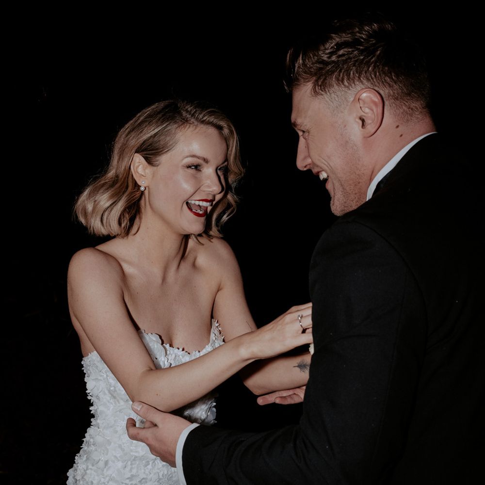 Bride in 3d floral short wedding reception dress laughing with the groom in black tuxedo 