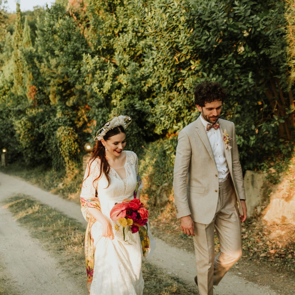 Bride and groom walk through greenery at italian destination wedding wearing white lace wedding dress, white floral structured bridal crown and greige suit