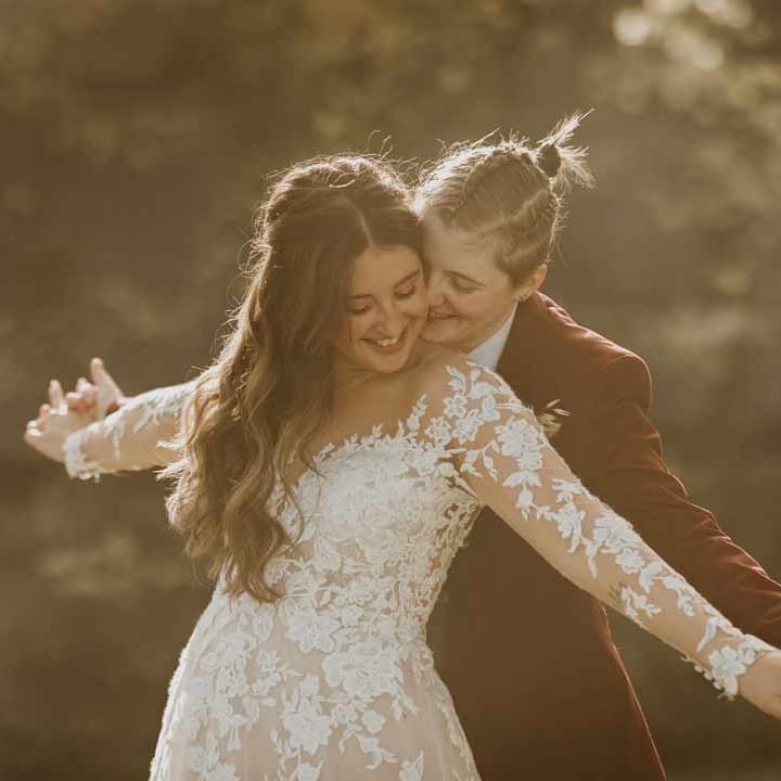 Bride in maroon bridal suit with grey waistcoat, dusky pink tie and autumnal dried flower boutonniere doing the Titanic pose with bride in long sleeve lace illusion sleeve wedding dress with overlay 