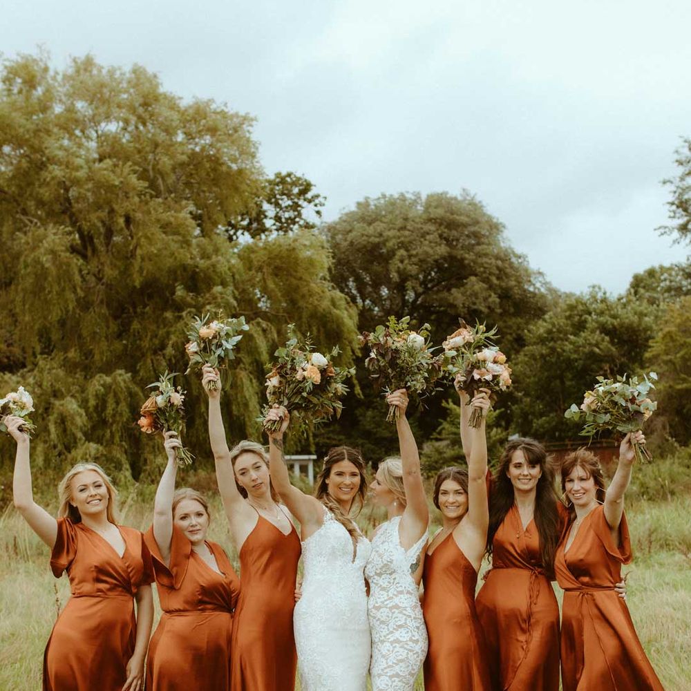 Two brides in white wedding dresses stand holding up bouquets with bridesmaids in burnt orange satin bridesmaid dresses 