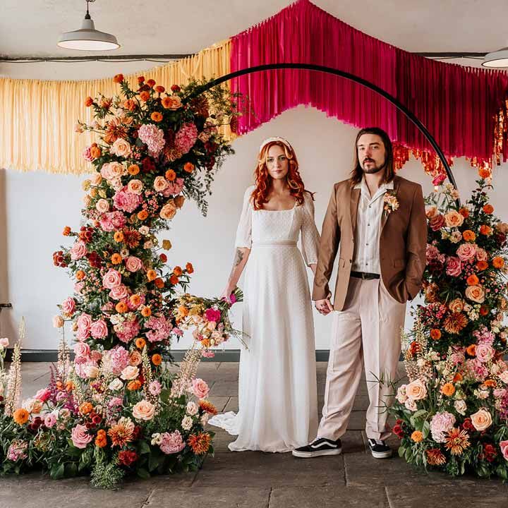 Groom in brown blazer, white shirt, grey suit trousers and vans shoes holding hands with bride in three quarter length puff sheer sleeve wedding dress with delicate polka dot overlay and square neck standing underneath circular floral arch with vibrant flowers and foliage attached and pink, red, yellow and metallic wedding streamers behind at Northern Monk Refectory Leeds
