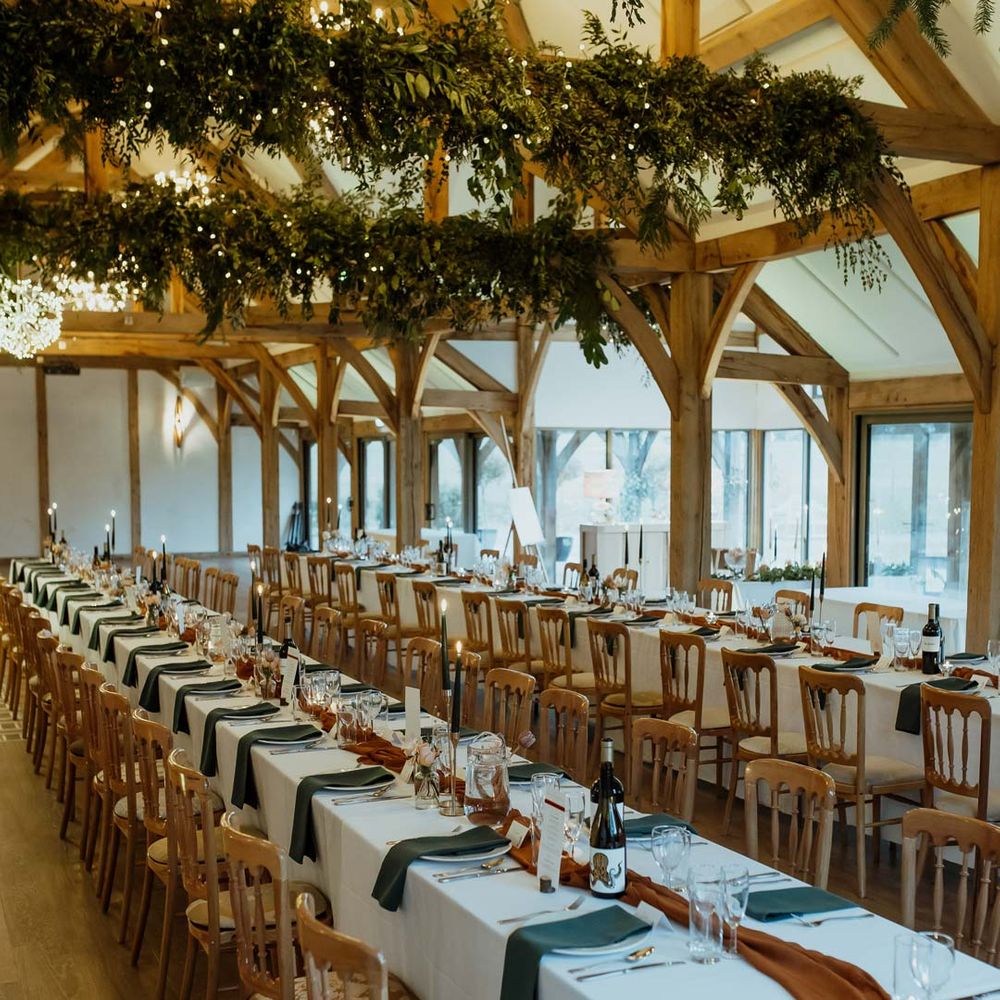 Classic wedding tablescape with white tablecloth, burnt orange wedding tablerunner, forest green wedding napkins, forest green tapered candles and bottles of wine with suspended foliage and fairy lights decor at wedding
