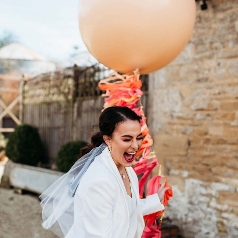 Bride in white bridal suit with closed toe bright green wedding heels and short bridal veil laughing outside Merriscourt wedding venue holding large balloon with colourful wedding streamers attached