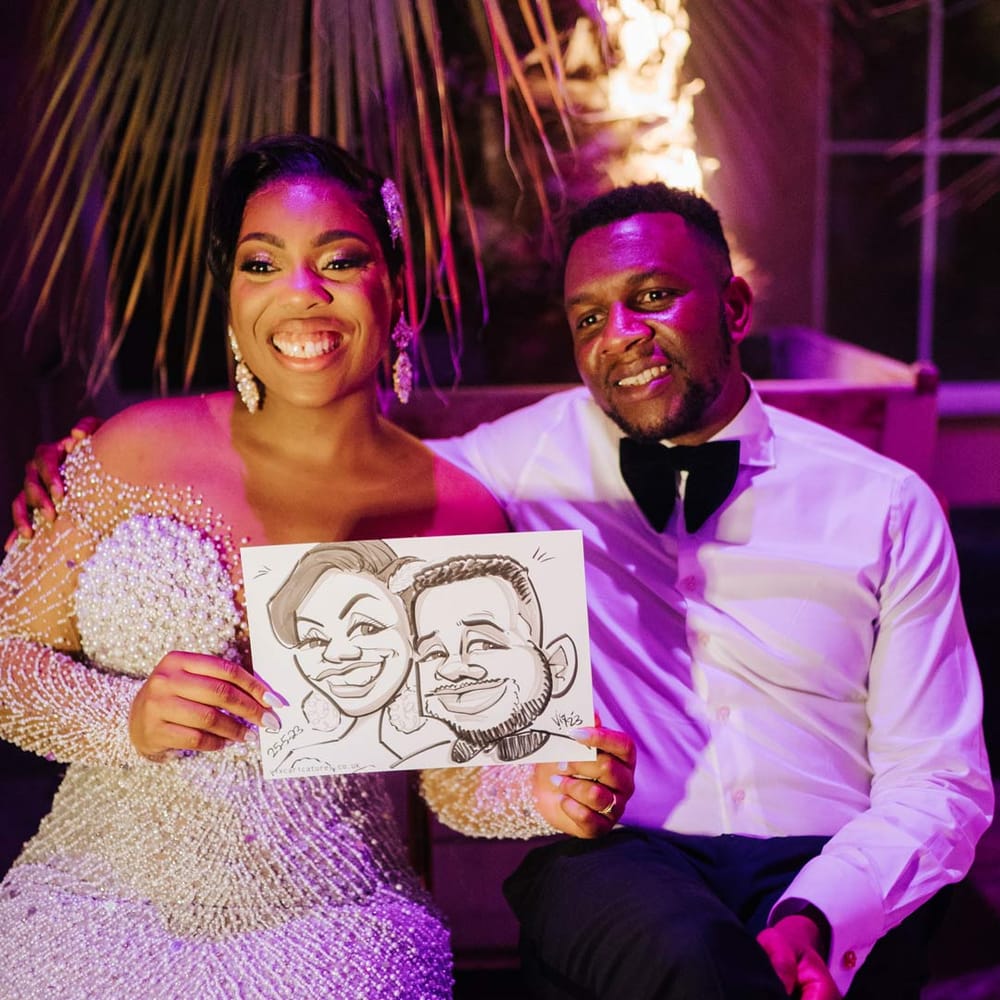 Groom in white shirt and velvet bowtie sitting with bride in illusion v neck sheer off shoulder long sleeve pearl embellished wedding gown holding up live wedding caricature painting
