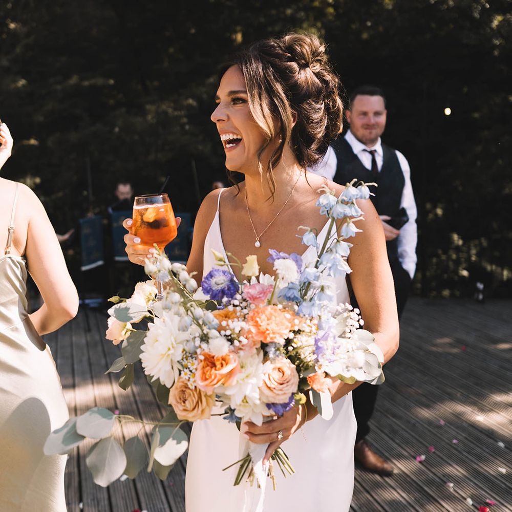Bride wearing satin slip wedding dress holding pastel wedding bouquet drinking champagne 