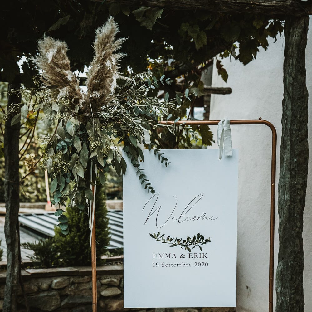 Wedding welcome sign hanging from a copper frame decorated in foliage and pampas grass flowers 