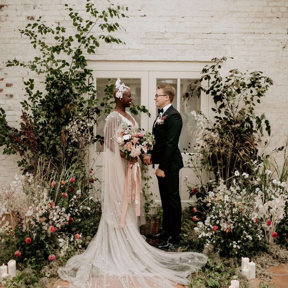 Black bride with alopecia standing at the altar in a blush pink Julita Bridal London wedding dress with cape with her groom in a tuxedo surrounded by wildflower arrangements 