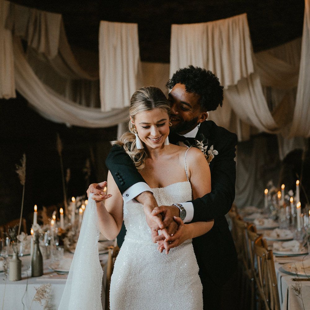 Groom in a tuxedo embracing his bride in a sparkly wedding dress as they stand inside their modern luxury wedding reception at Pentney Abbey, Norfolk