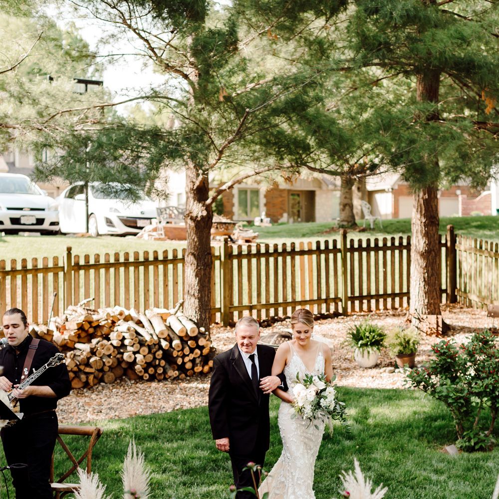 Bride in white Made With Love wedding dress walks up the pampas grass lined aisle in garden arm in arm with father in black suit for backyard wedding