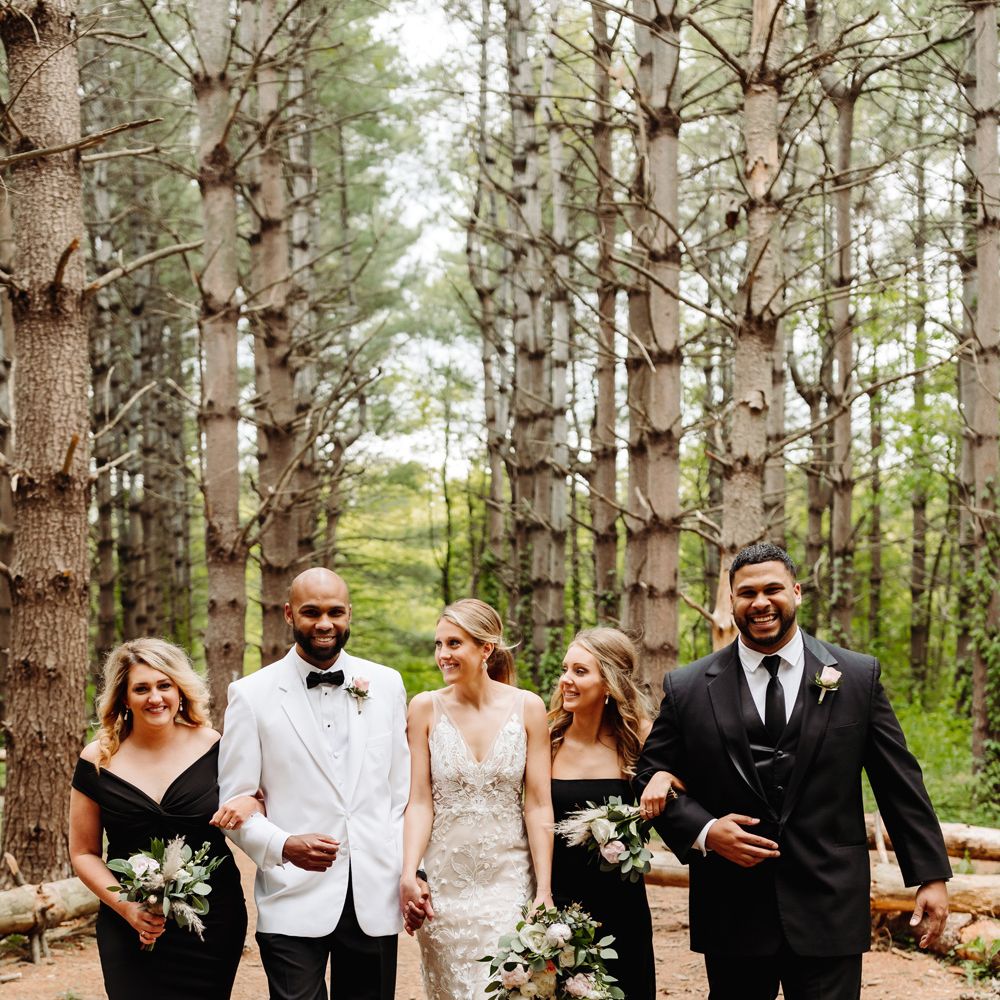 Bride in white embroidered Made With Love wedding dress holds hands with groom in white tuxedo jacket as they walk through woodland with two bridesmaids in black dressed and groomsman in black suit after backyard wedding