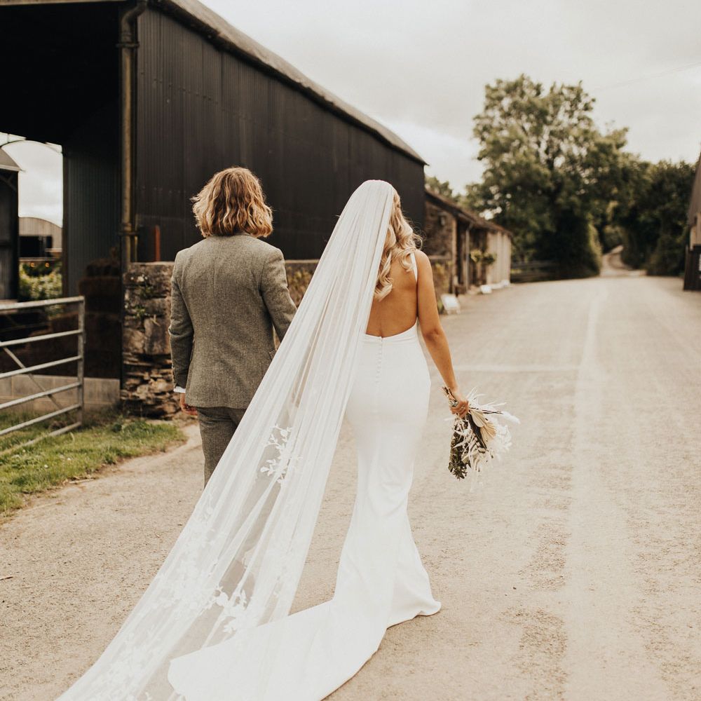 Bride in white open back Made With Love wedding dress with train and long embroidered veil holds white and green bridal bouquet and hand of groom in sage green suit in the other as they walk down country lane after wedding ceremony at Anran Devon