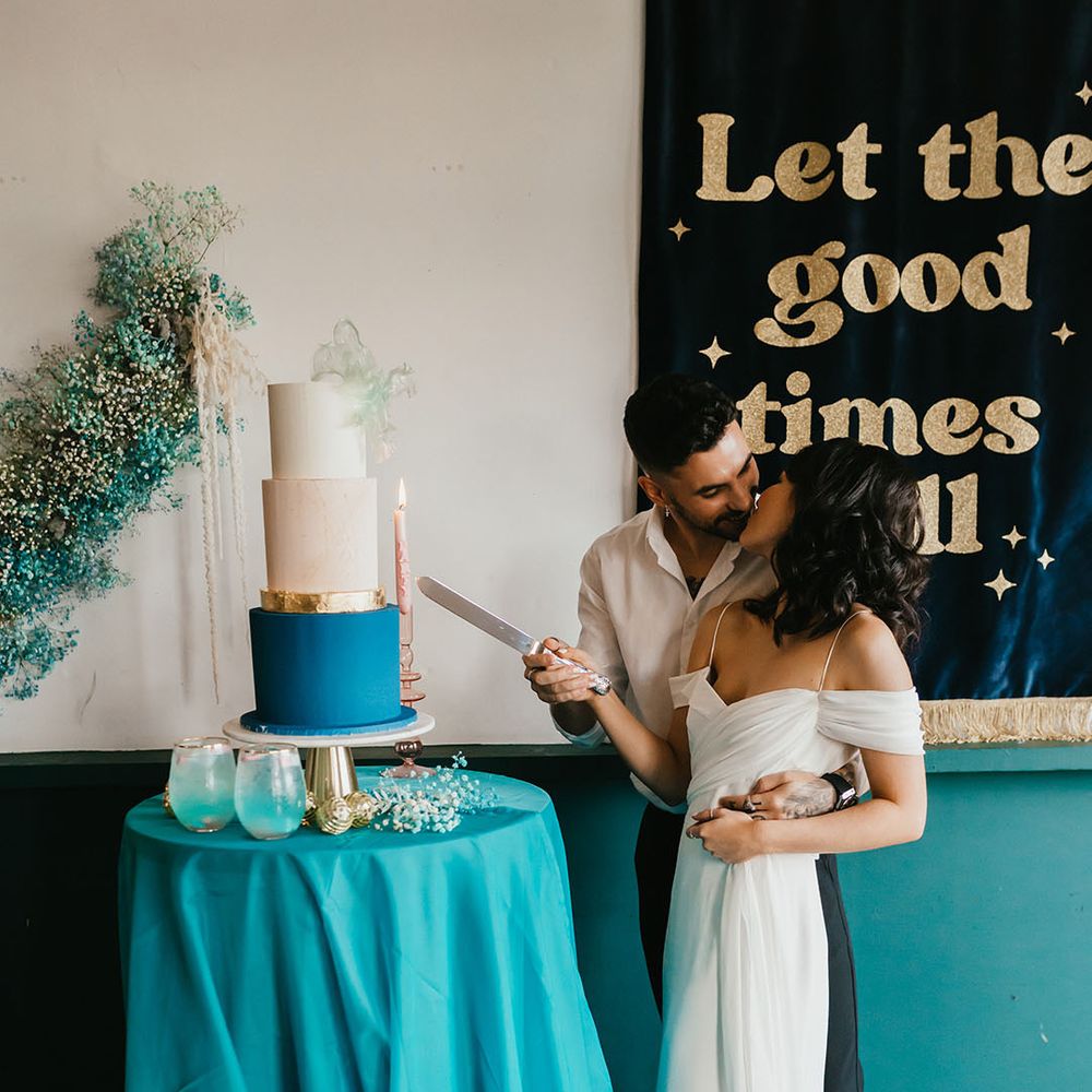 Groom in an open collar white shirt kissing his bride in a cold shoulder wedding dress as they cut their contemporary wedding cake 