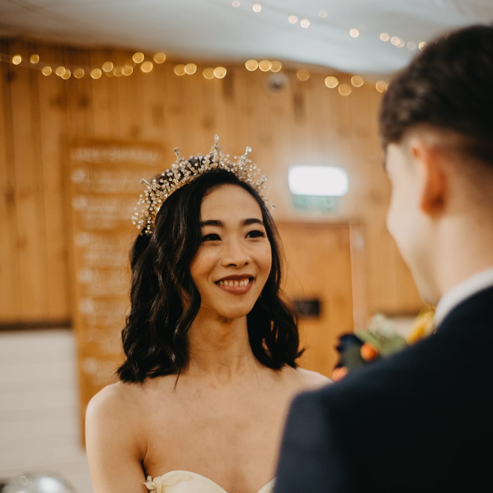 Smiling bride in strapless feathered wedding dress and bridal headband smiles at groom during rustic wedding ceremony at The Wellbeing Farm in Lancashire