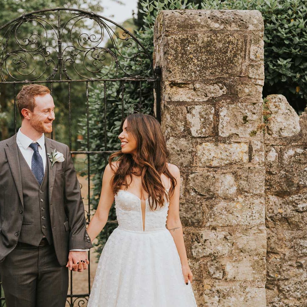 Groom in grey three piece suit, blue tie and white rose buttonhole smiles whilst holding hands with bride in cami strap Pronovias wedding dress as they stand by walled garden in Buckinghamshire