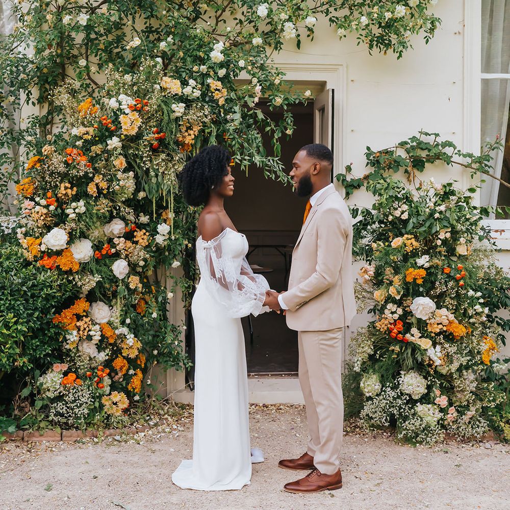 Bride in a strapless wedding dress with sheer sleeves and groom in beige suit holding hands next to the vine at Modern Hall, London with orange flowers