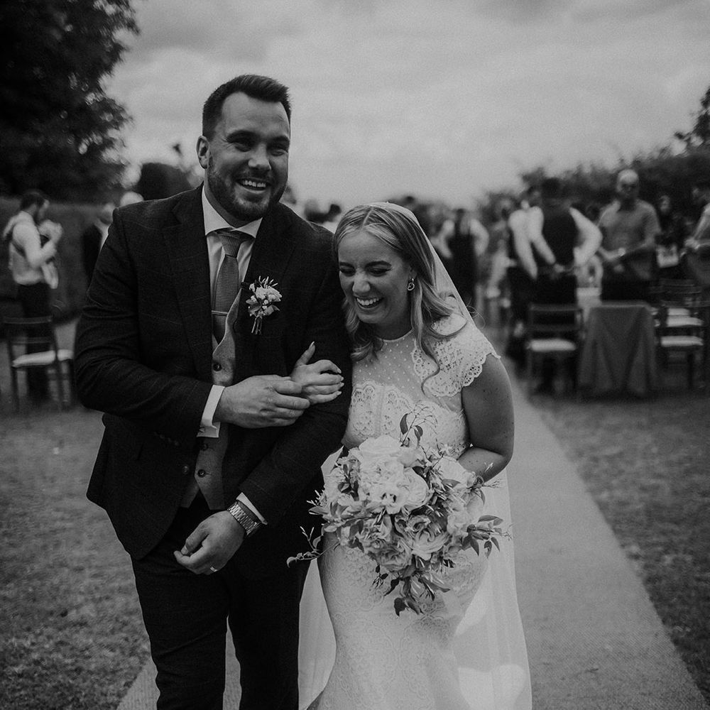 Black and white portrait of the bride in a lace wedding dress arm in arm with her groom walking up the aisle 