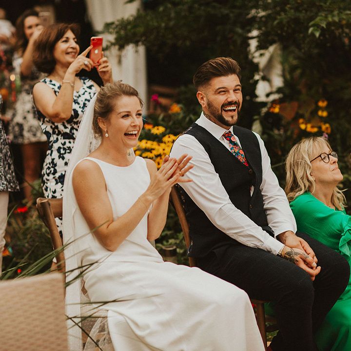 Bride in a slip wedding dress and pinned up hair and groom in a navy waistcoat and floral tie sitting on wooden chairs laughing during the speeches 