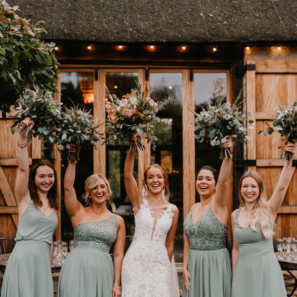 Bride in deep v lace wedding dress with tulle skirt and bridesmaids in sage green bridesmaid dresses hold up mixed wedding bouquets outside barn after wedding ceremony