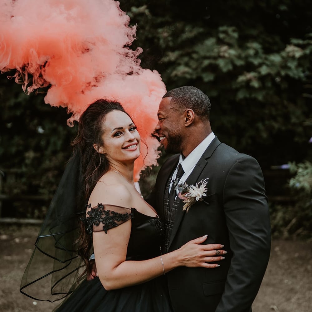 Bride & groom hold pink smoke bomb as they embrace after wedding ceremony