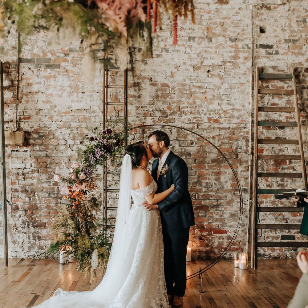 Bride in lace wedding dress and veil kisses groom in blue check suit in front of boho floral moongate with pink, purple and green florals for wedding ceremony in industrial venue