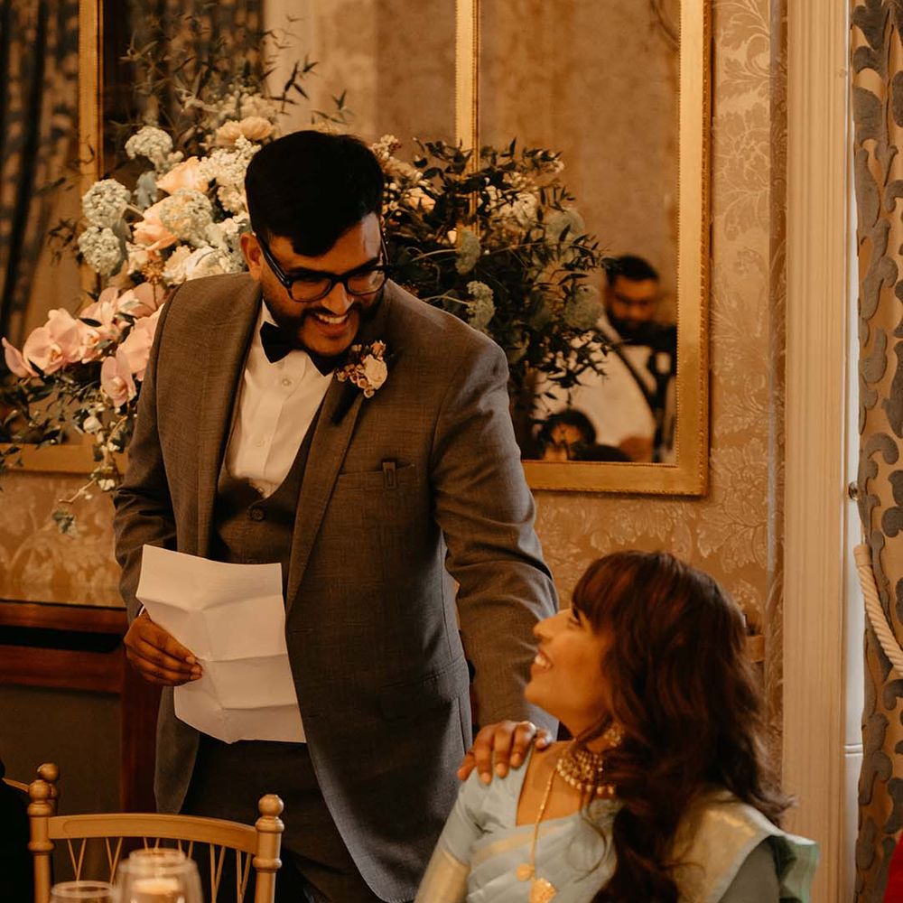 Bride in mint green saree with golden details and groom giving speech in dark grey suit black bow tie and boutonniere 