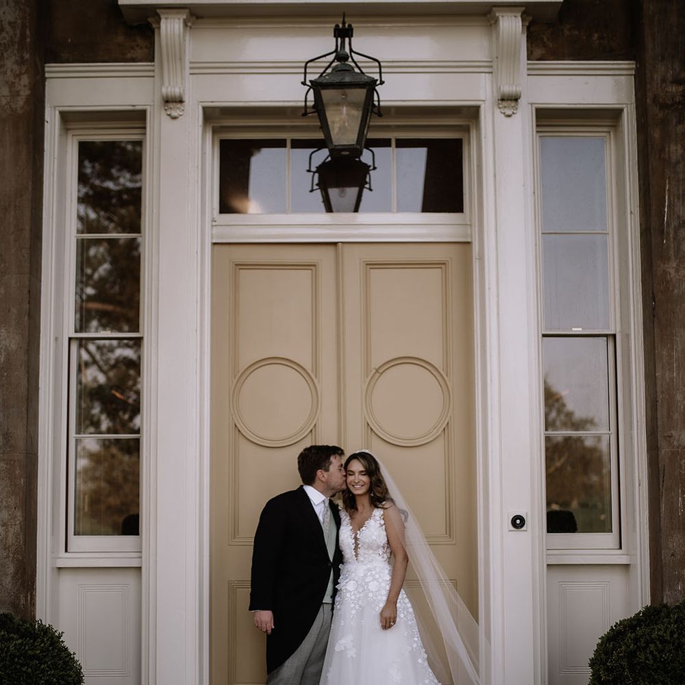 Groom in morning suit kisses bride on the cheek in Mira Zwillinger wedding dress in front of large doorway 