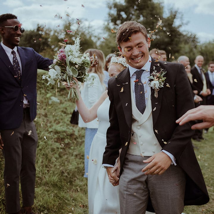 Groom walks through colourful confetti and scrunches his face and holds hands with the bride 