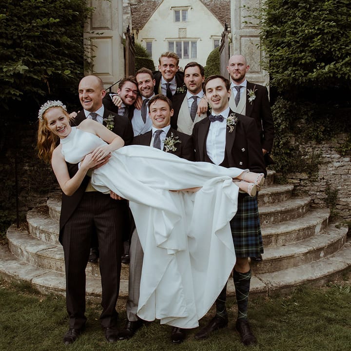 Groom and groomsmen lift the bride as they all pose together in their matching morning suits 
