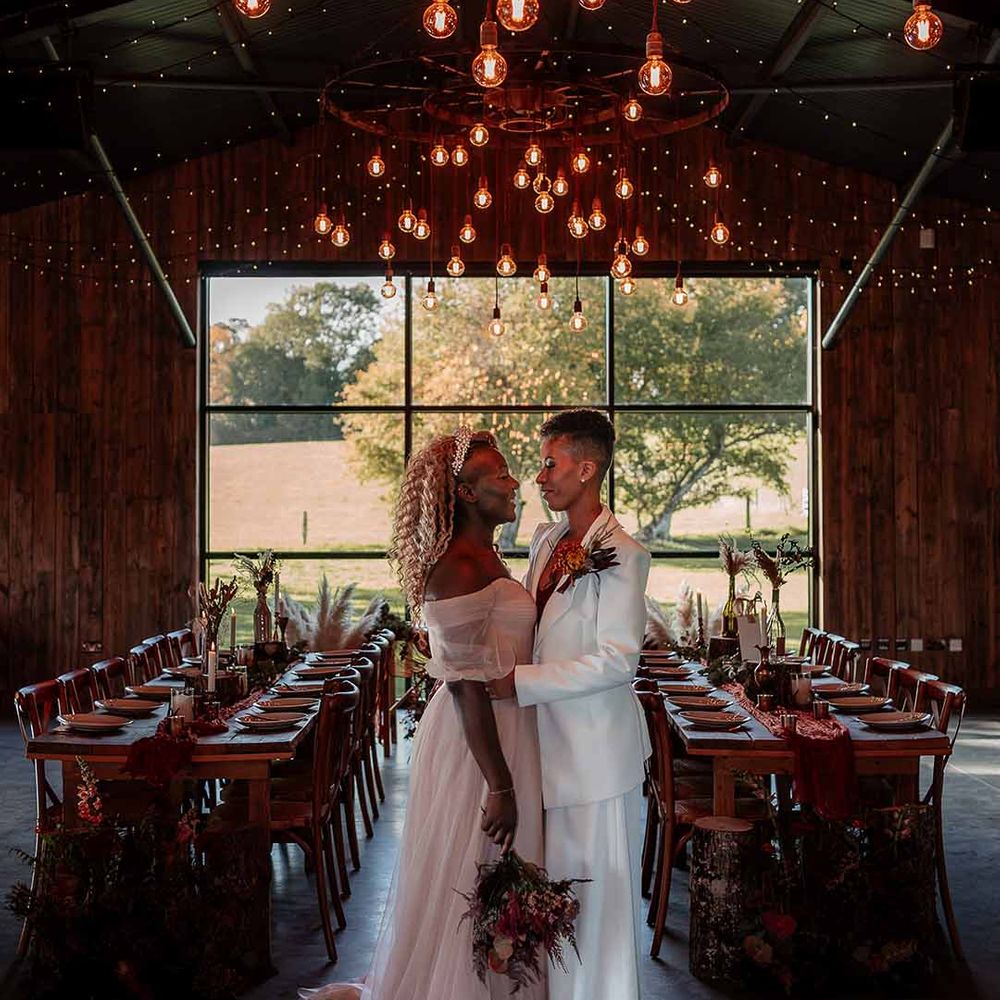 Brides in complimenting ethereal boho bridal outfits embracing with large autumnal dried flower bouquet standing underneath festoon chandelier lighting and fairy lights
