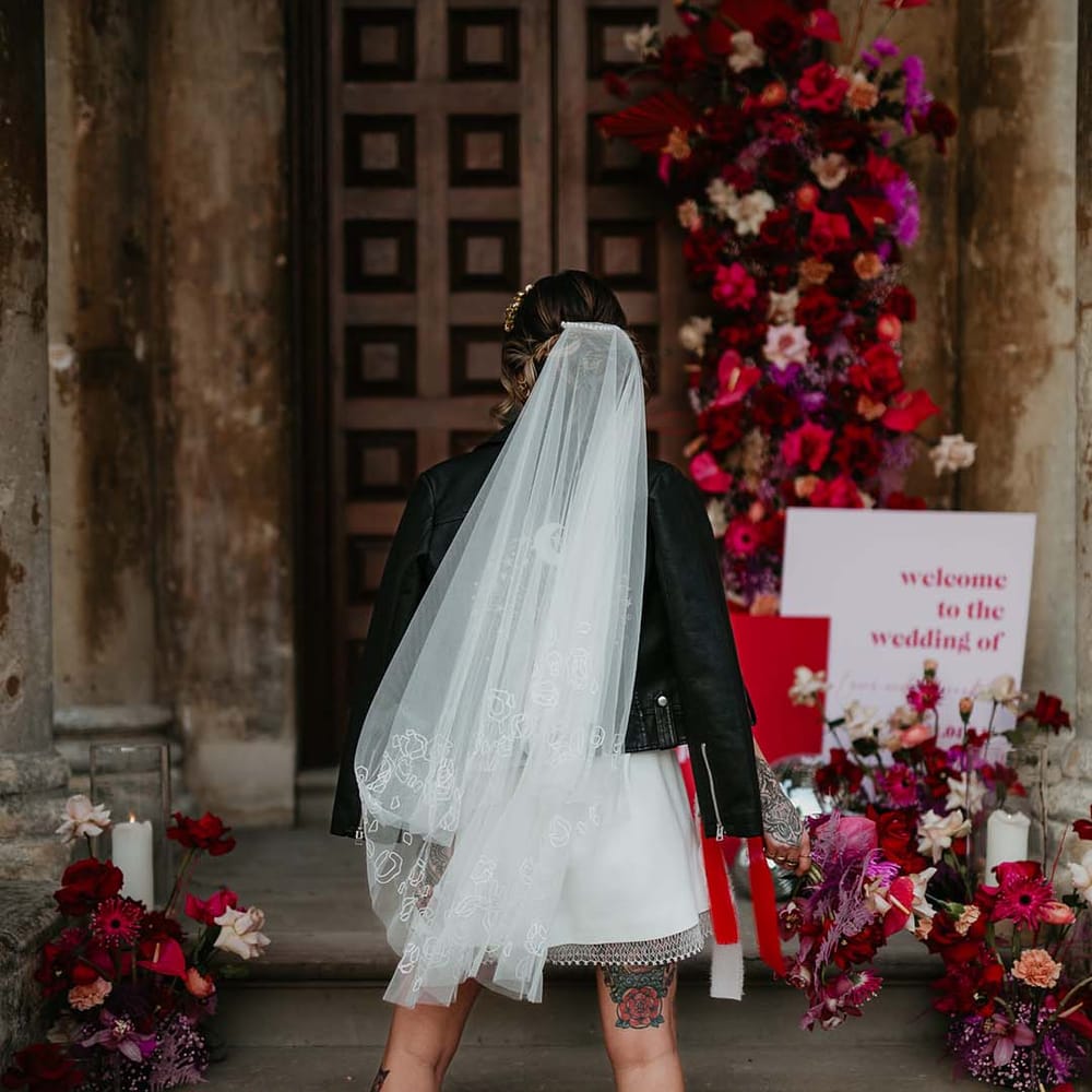 Bride in short wedding veil with delicate flower embroidery at the base and white patent heeled ankle boots with black soles surrounded by hot pink, bright pink and viva magenta dahlias, orchids, roses, amaryllis, anthuriums and dried flowers 