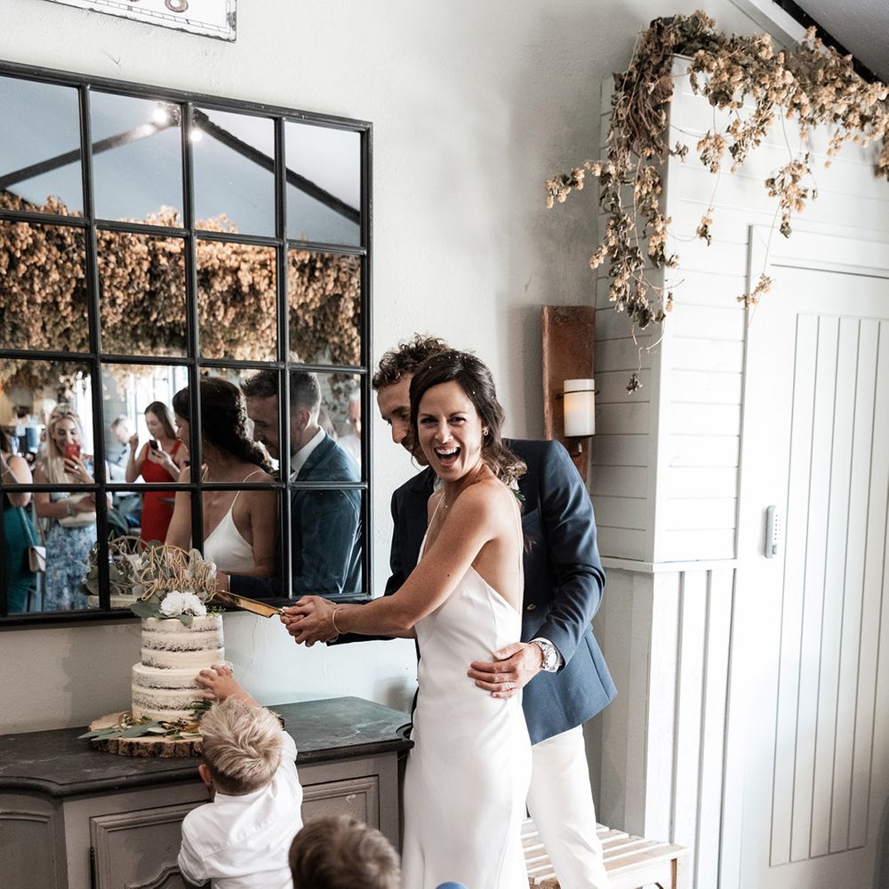 Bride cuts two tier naked wedding cake with her groom and children at Gate Street Barn