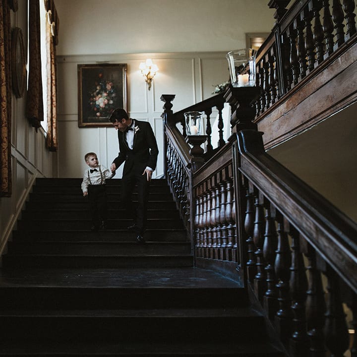 Tiny wedding guest wearing a white shirt, braces and black bow tie walks with grown up in black tie 