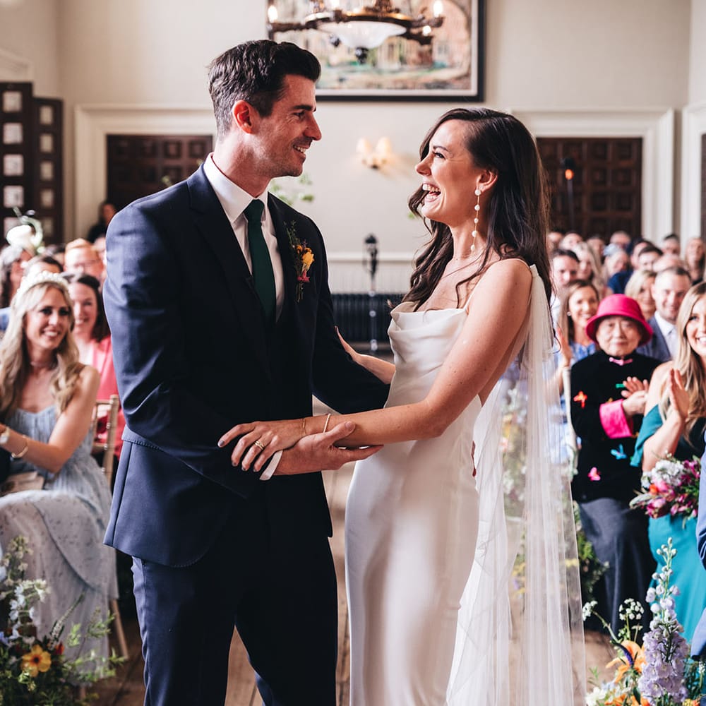 The bride in a sleek cowl neck satin wedding dress with long pearl earrings laughs at the altar with the groom in a navy suit 
