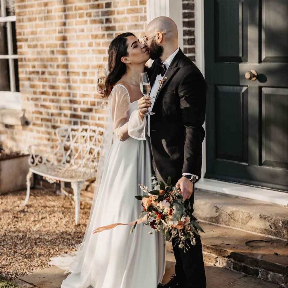 Bride in square neck wedding dress with sheer puff sleeves holding a glass of champagne sharing a kiss with groom in classic black tuxedo with black bowtie 
