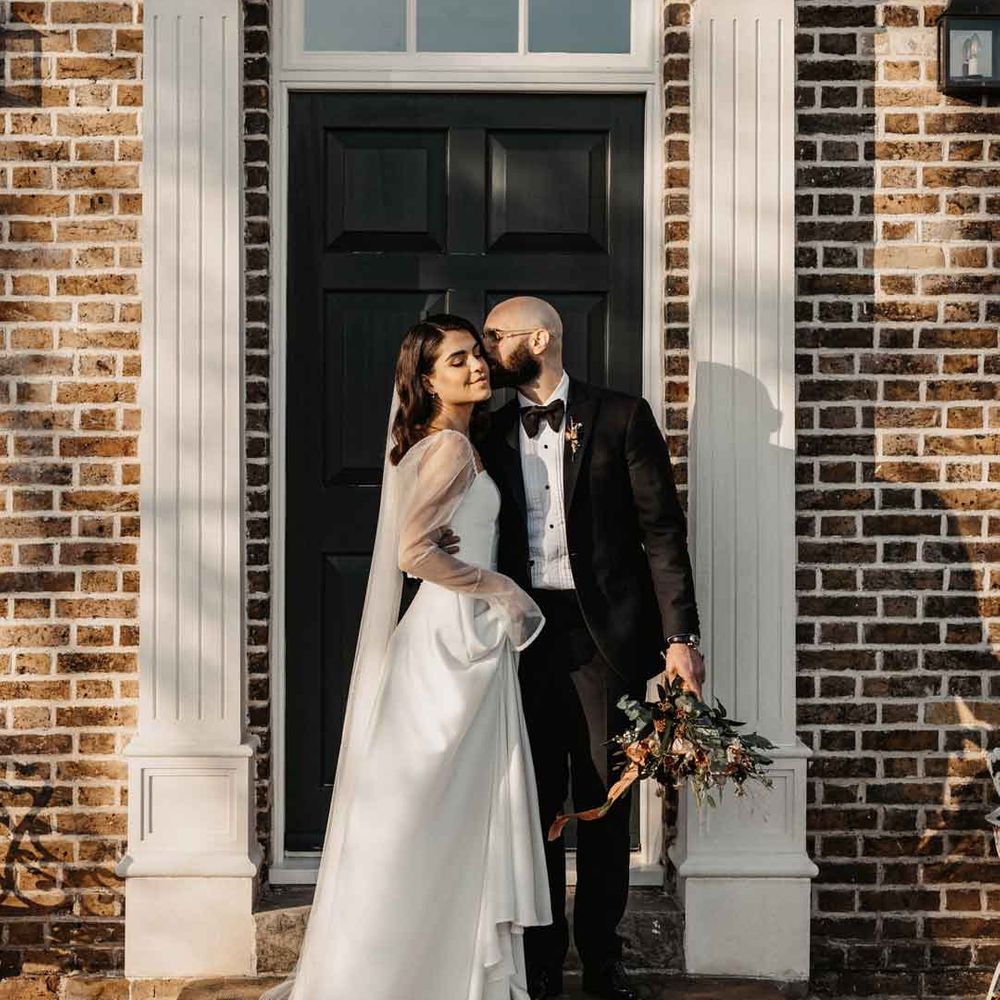 Groom in classic black tuxedo with black bowtie embracing bride in square neck wedding dress with sheer puff sleeves at Elmley Nature Reserve wedding venue 