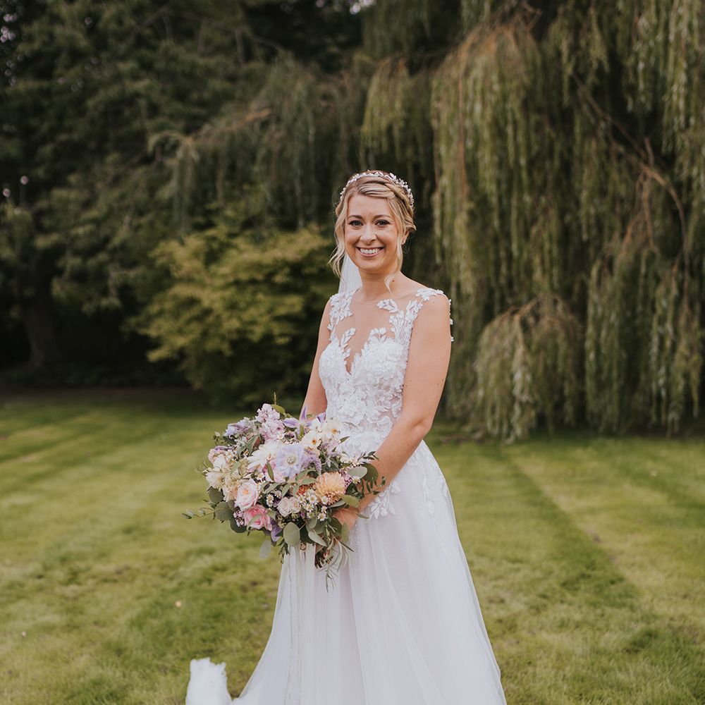 Dando London wedding dress with illusion lace and tulle skirt holding a pastel wedding bouquet with her blonde hair in a braided hairstyle and sparkly headpiece 