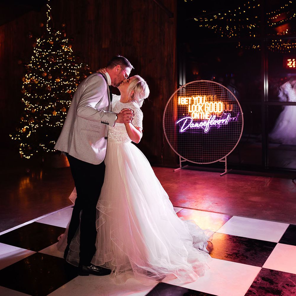 The bride in a tulle bridal skirt dances with the groom on a checkered dance floor with festoon lighting and Christmas tree decoration with neon sign