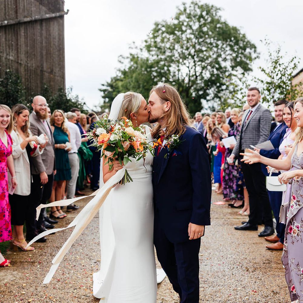 Bride holding a colourful wedding bouquet sharing a kiss with the groom at the end of their confetti exit 