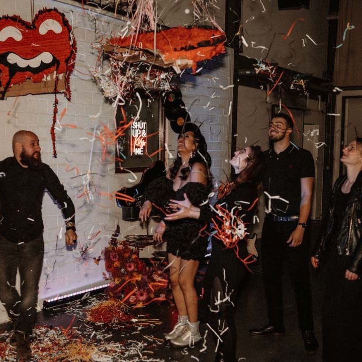 Bride in black ruffle off shoulder reception dress and red drip effect sunglasses with groom in black shirt and black drip effect sunglasses standing with wedding guests batting the Rolling Stones wedding piñata 