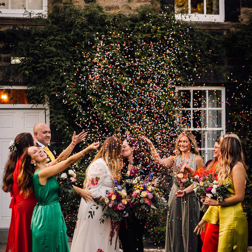 Bridesmaids in mismatched bridesmaid dresses in bold colours throw confetti over the two brides 