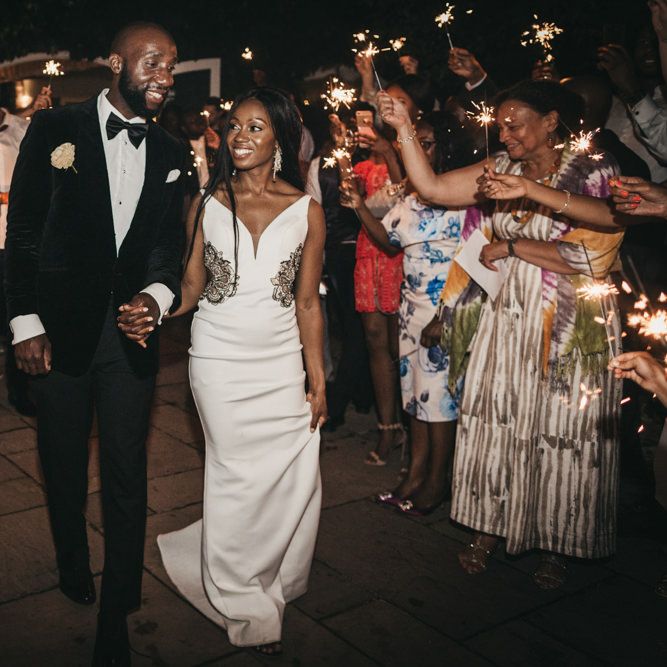 Sparkler moment with the Black bride in a fitted wedding dress with waist detail holding her grooms hand in a black tuxedo 