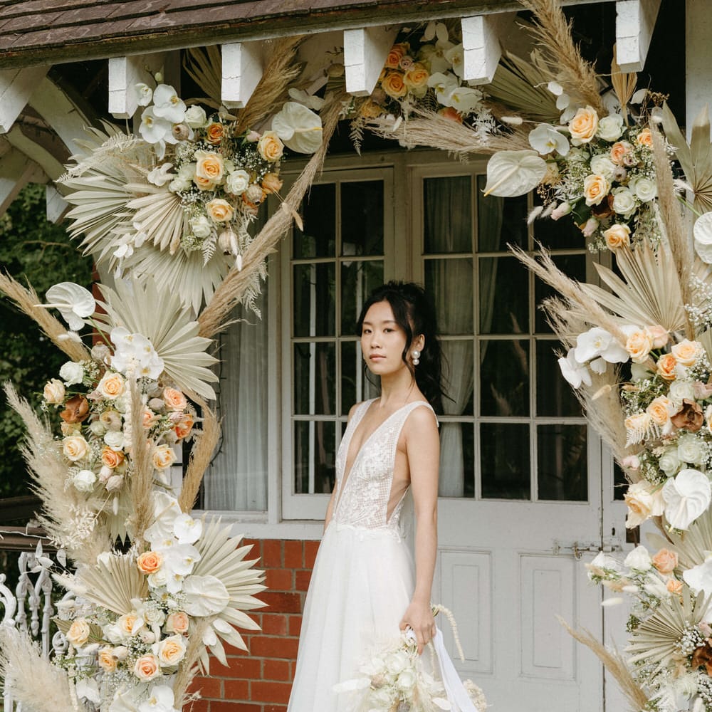 Bride at Berwick lodge wearing a plunge neck wedding gown with tulle skirt, under an arch of dried palms, pampas grass, white anthuriums, roses and orchids