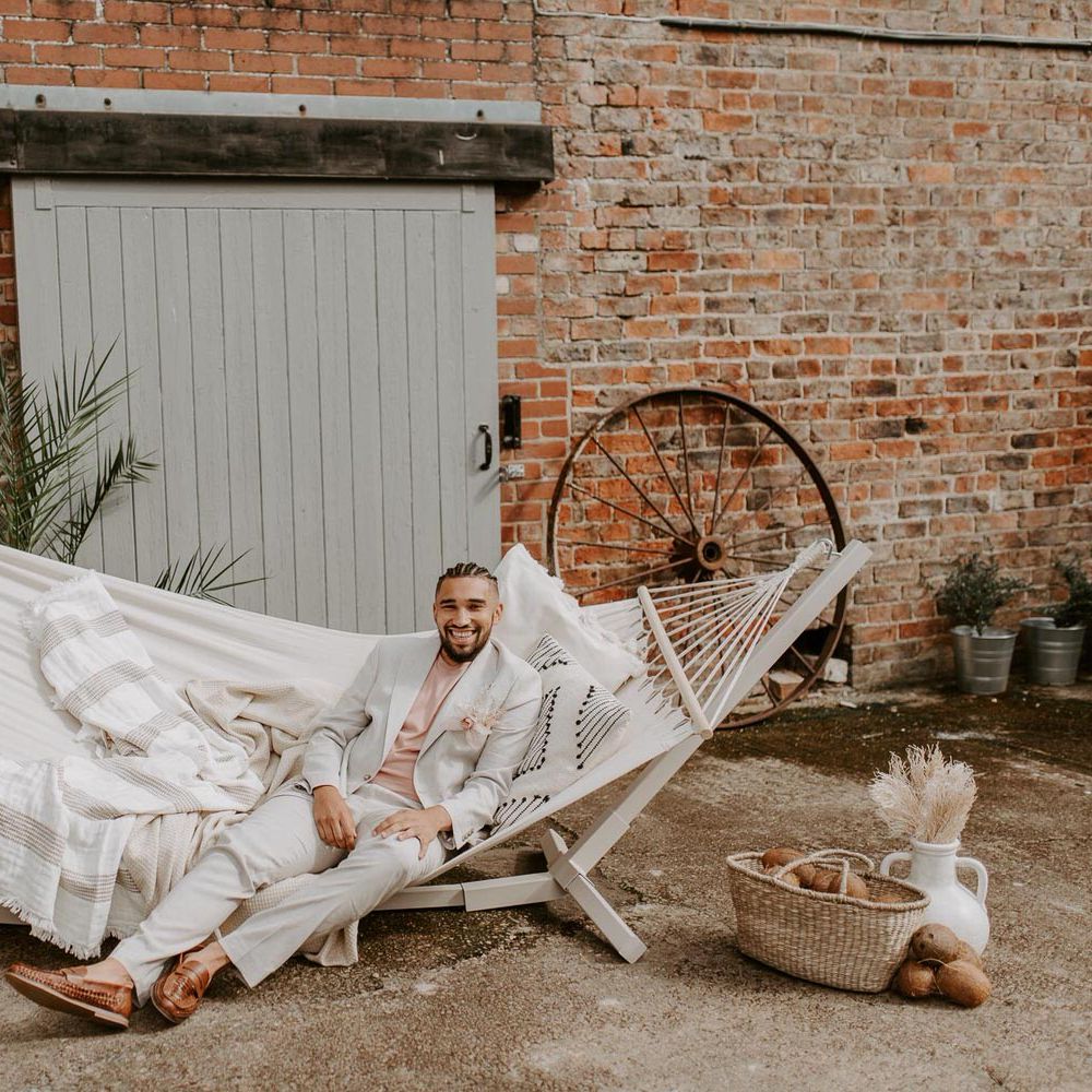 Groom in cream suit sitting on a large hammock for tropical wedding theme