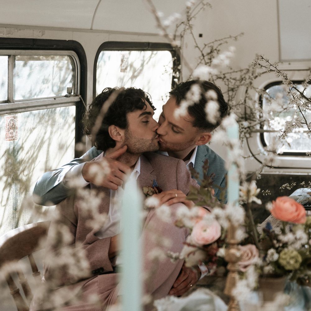Grooms kiss whilst sat at rustic wooden table with pastel floral bouquets 