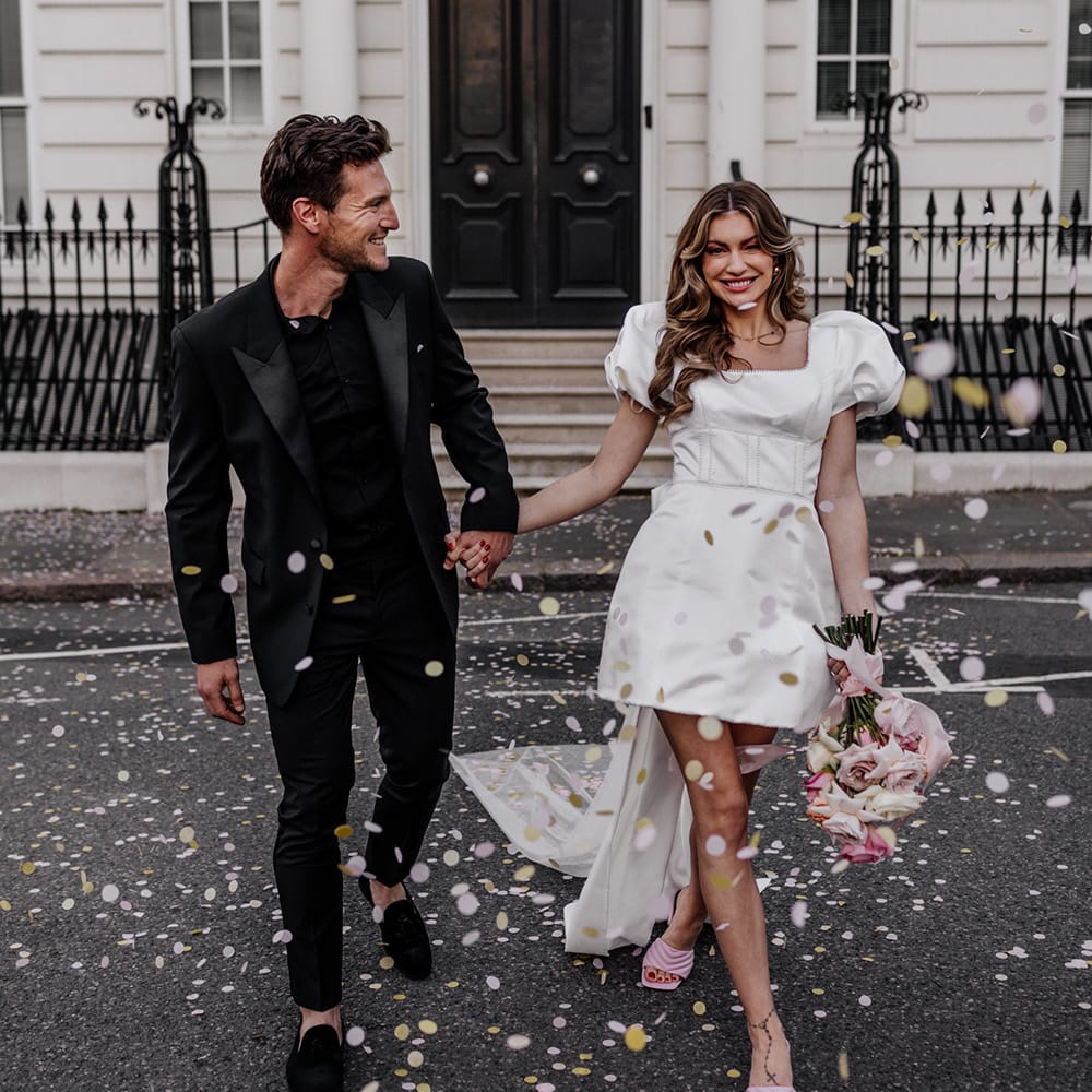 Confetti moment in the streets of London with bride in a knee length wedding dress with training bow back detail and groom in an all black suit 