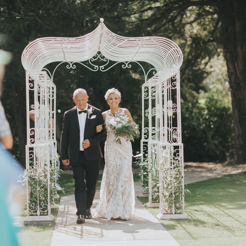 A bride in a lace wedding dress carries a bouquet and is linked arms with a man in a black tuxedo. They walk down an outdoor aisle.